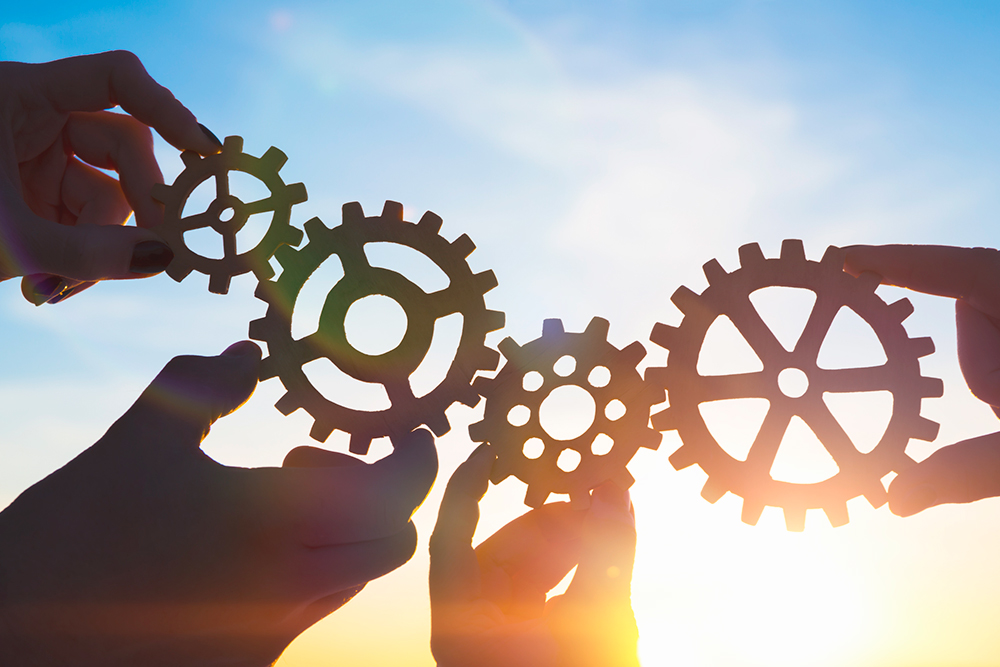 Four businessmen's hands assembling gears and puzzle pieces, symbolizing teamwork and collaboration.