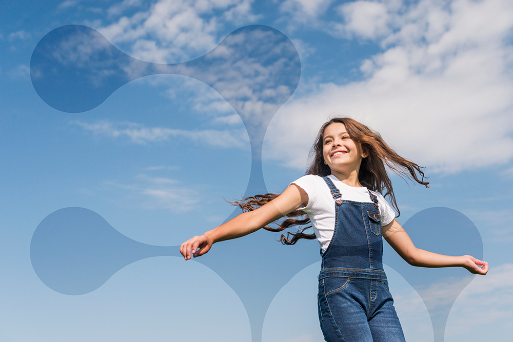 Smiling young girl with long hair wearing denim overalls and a white T-shirt, playing outdoors under a blue sky with clouds.