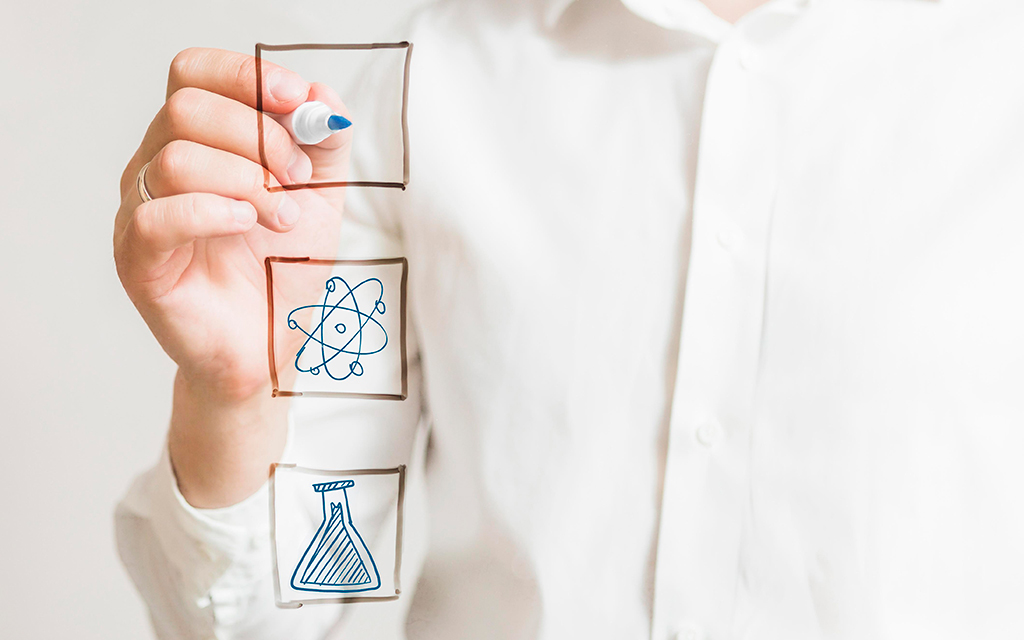 Person drawing science icons, including an atom and a laboratory flask, on a transparent glass surface.